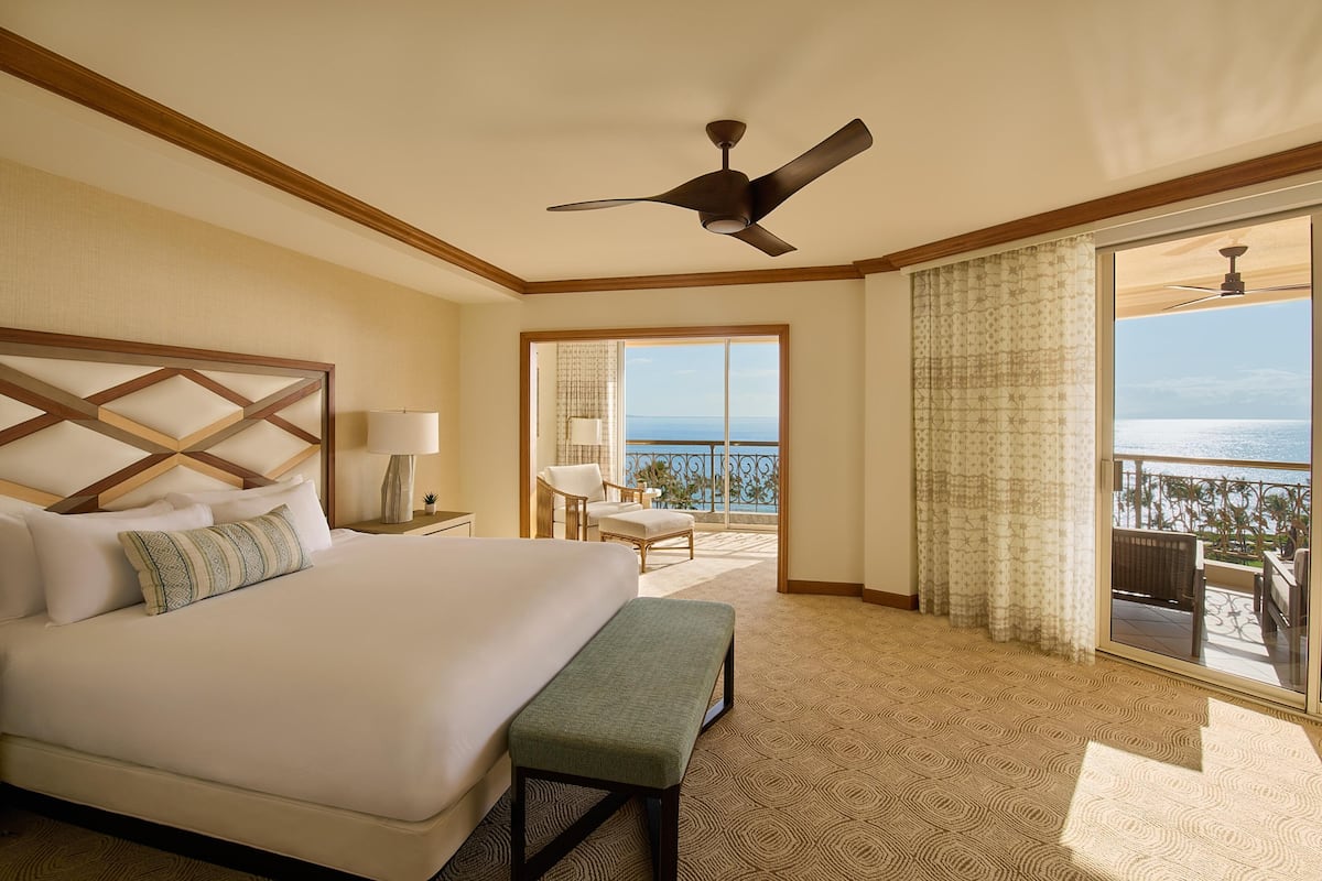 A hotel room with a large bed, a ceiling fan, a bench, a nightstand, a lamp, a view of the ocean through a balcony door, and a patterned carpet.