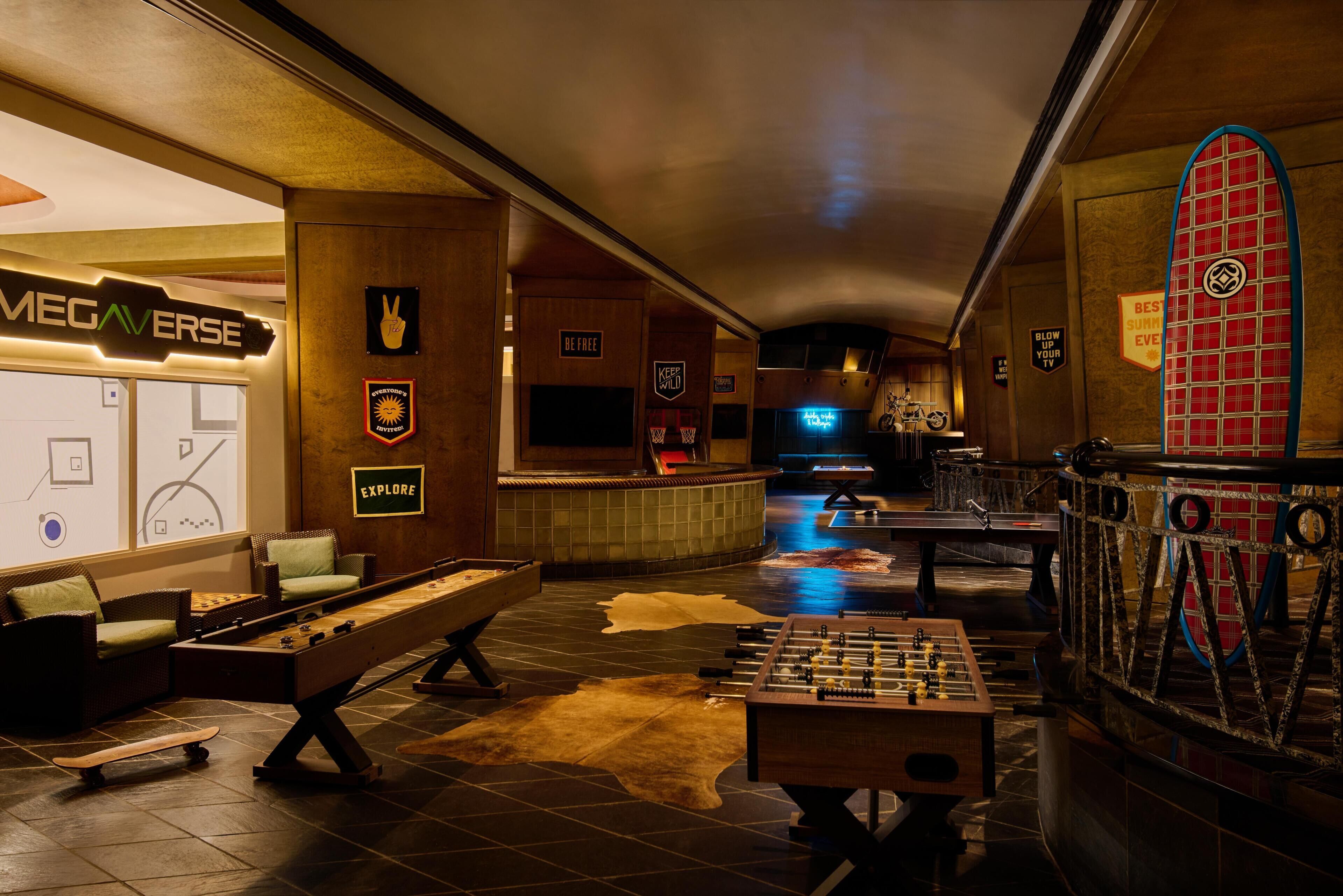 A room with a foosball table, a pool table, and a bar area with a neon sign.