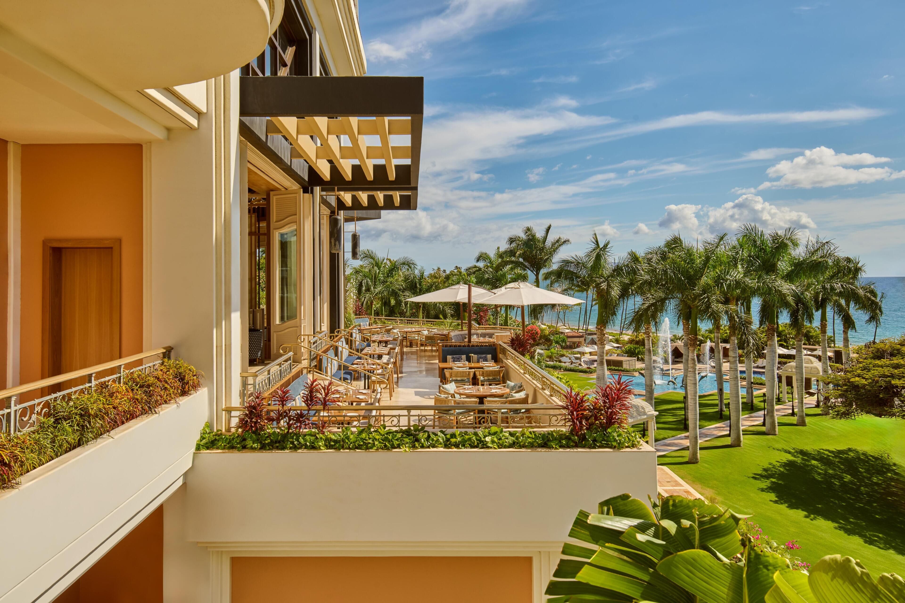 Un balcón de hotel con mobiliario exterior, piscina y palmeras.
