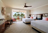 A hotel room with two beds, a ceiling fan, a desk with a chair, a window with a view of palm trees, and a patterned carpet.