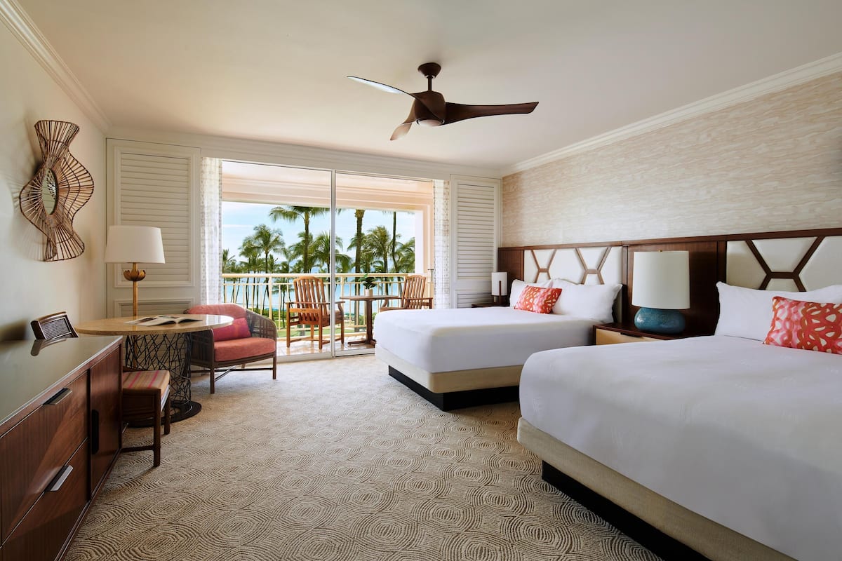 A hotel room with two beds, a ceiling fan, a desk with a chair, a window with a view of palm trees, and a patterned carpet.