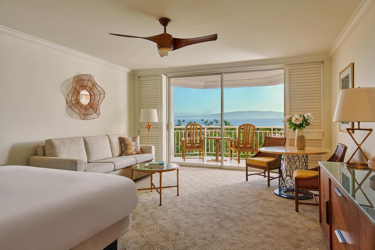 A hotel room with a bed, a sofa, a dining table with chairs, a ceiling fan, and a view of the outdoors.