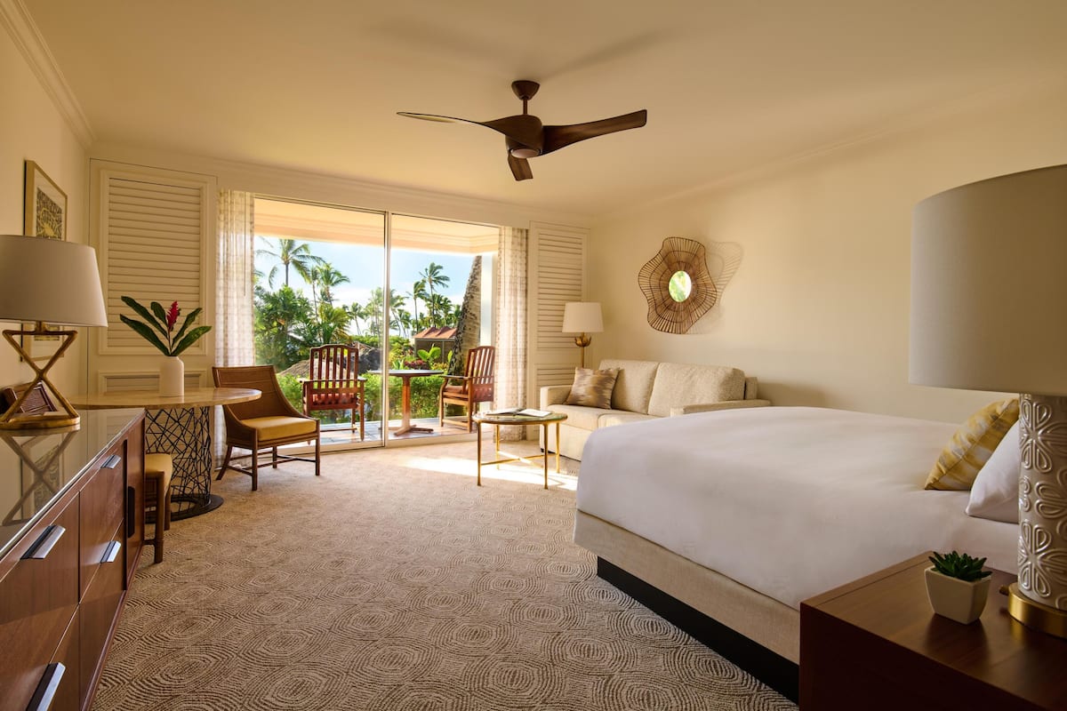 A hotel room with a bed, a desk, a chair, a table, and a balcony with a view of palm trees.