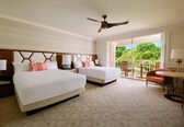 A hotel room with two beds, a ceiling fan, a balcony with a table and chairs, and a view of greenery.