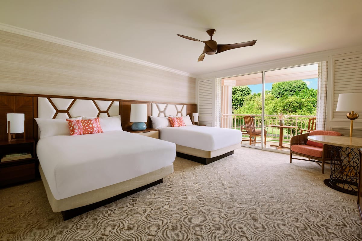 A hotel room with two beds, a ceiling fan, a balcony with a table and chairs, and a view of greenery.