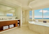 A modern bathroom with a large mirror, a walk-in shower, a bathtub, and a view of the ocean.