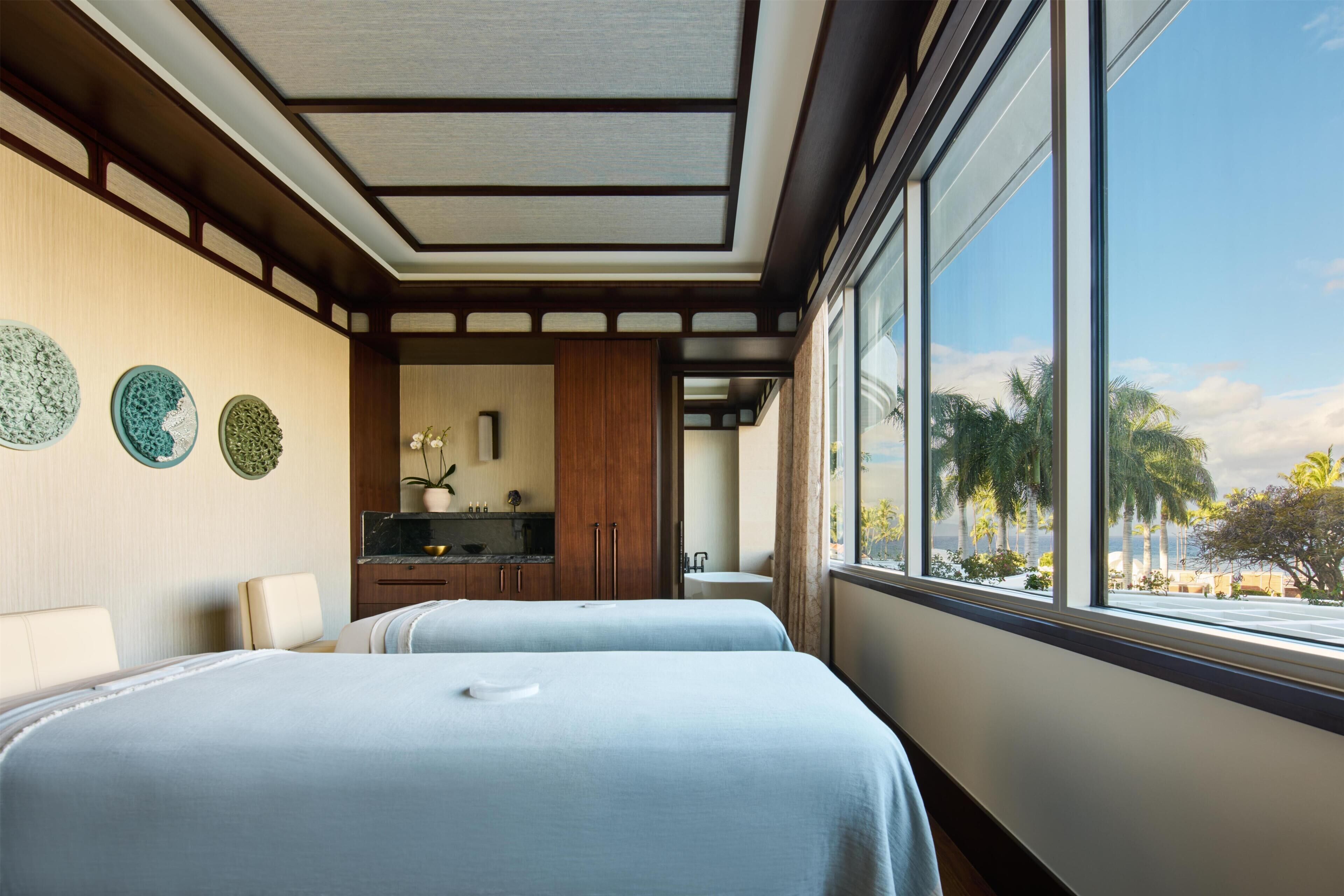 A hotel room with two beds, a kitchenette, and a large window with a view of palm trees.