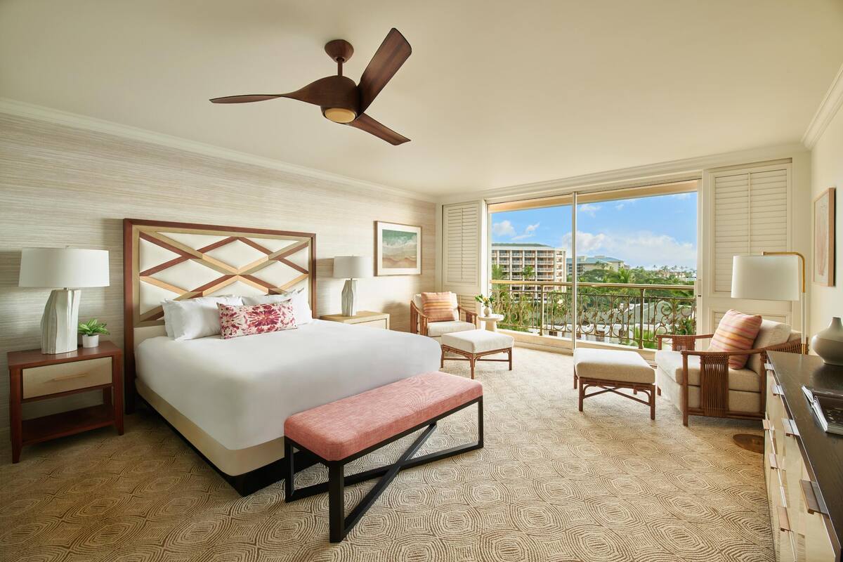 A hotel room with a large bed, a ceiling fan, a balcony with a view, and a desk with a lamp.