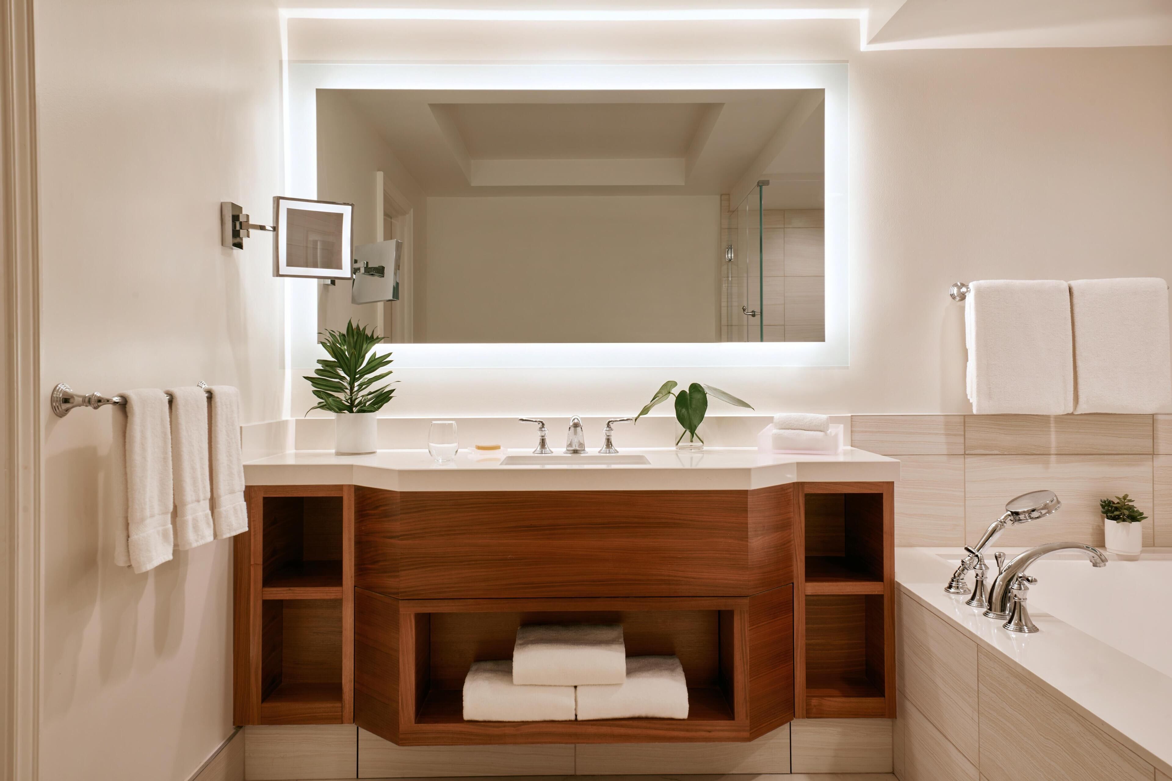 A modern bathroom with a large mirror, a wooden vanity, a bathtub, and a potted plant.