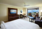 A hotel room with a large bed, a television on the wall, a desk with a chair, a small table, and a balcony with chairs.
