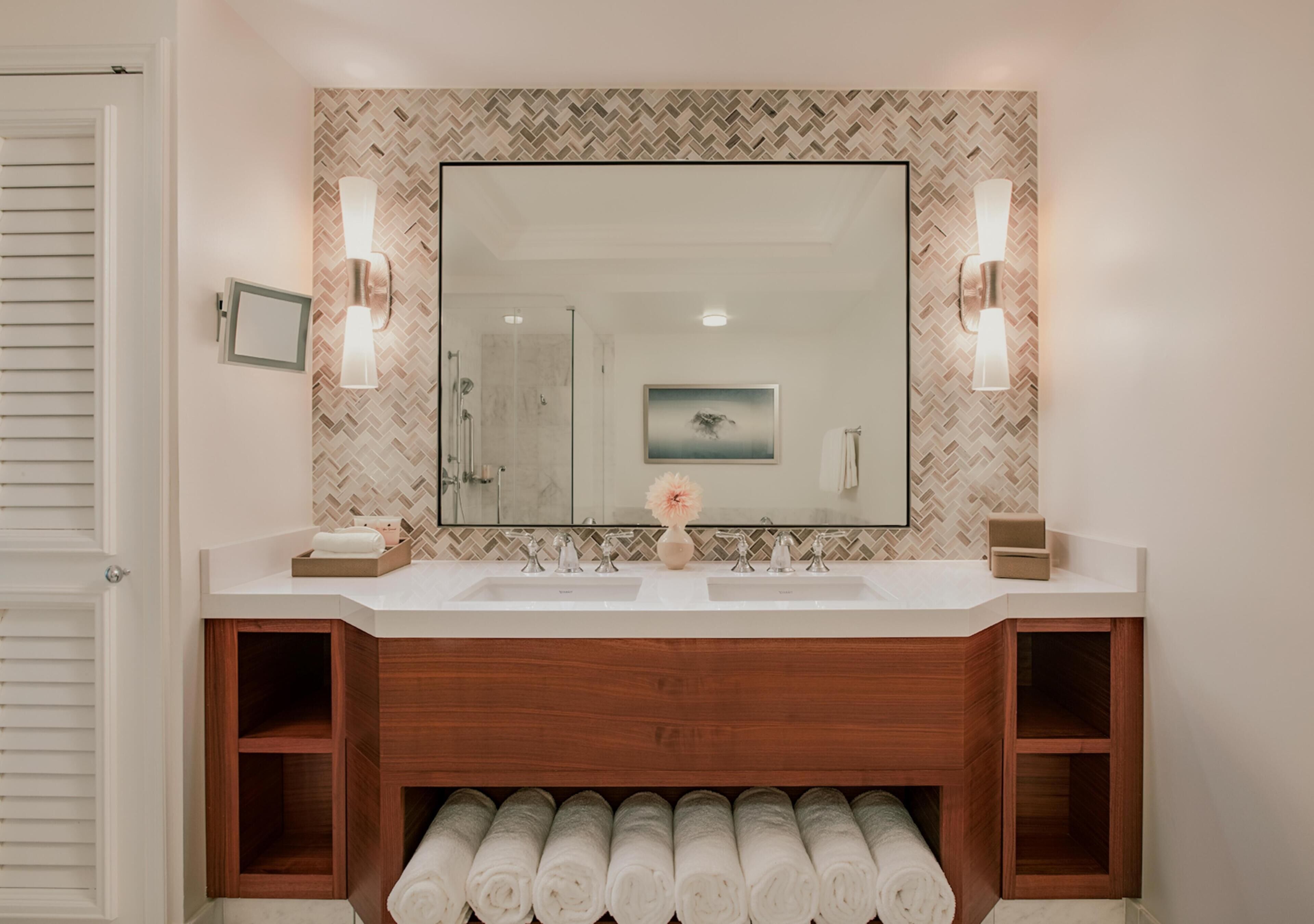A bathroom with a large mirror, two wall-mounted lights, a herringbone-patterned backsplash, and a wooden vanity with a towel rack below.