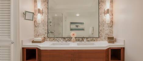 A bathroom with a large mirror, two wall-mounted lights, a herringbone-patterned backsplash, and a wooden vanity with a towel rack below.