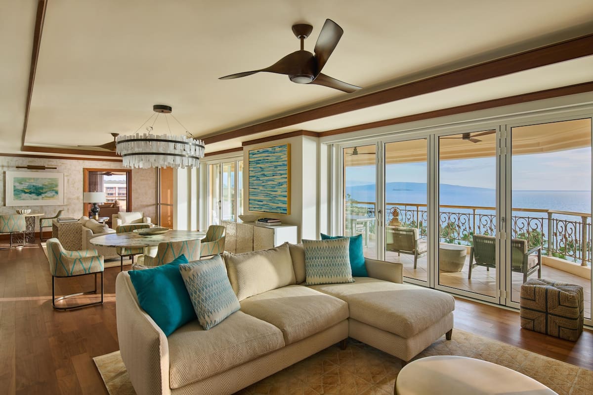 A spacious living area with a large sectional sofa, a chandelier, ceiling fans, and a balcony with a view.