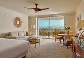 A hotel room with a bed, a sofa, a dining table with chairs, a ceiling fan, and a view of the outdoors.