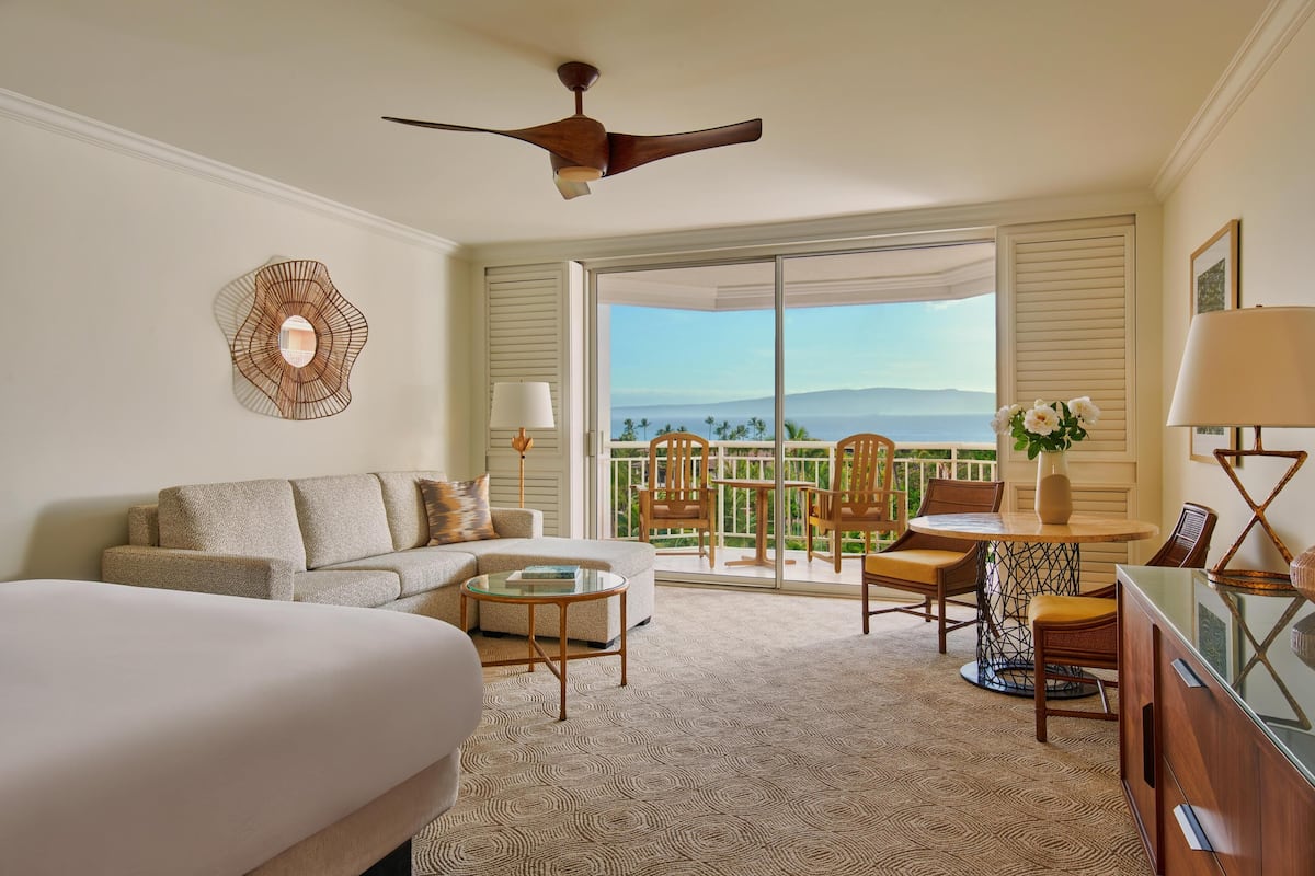 A hotel room with a bed, a sofa, a dining table with chairs, a ceiling fan, and a view of the outdoors.