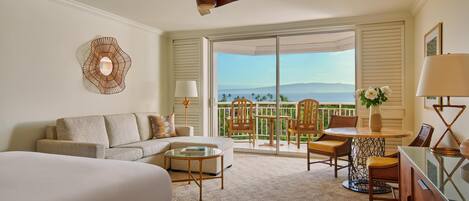 A hotel room with a bed, a sofa, a dining table with chairs, a ceiling fan, and a view of the outdoors.