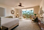 A hotel room with a bed, a desk, a chair, a dining table, and a balcony with a view of palm trees.