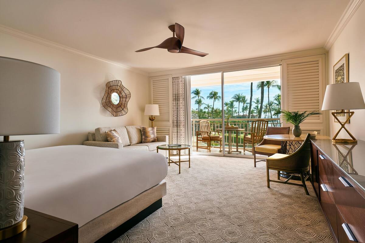 A hotel room with a bed, a desk, a chair, a dining table, and a balcony with a view of palm trees.