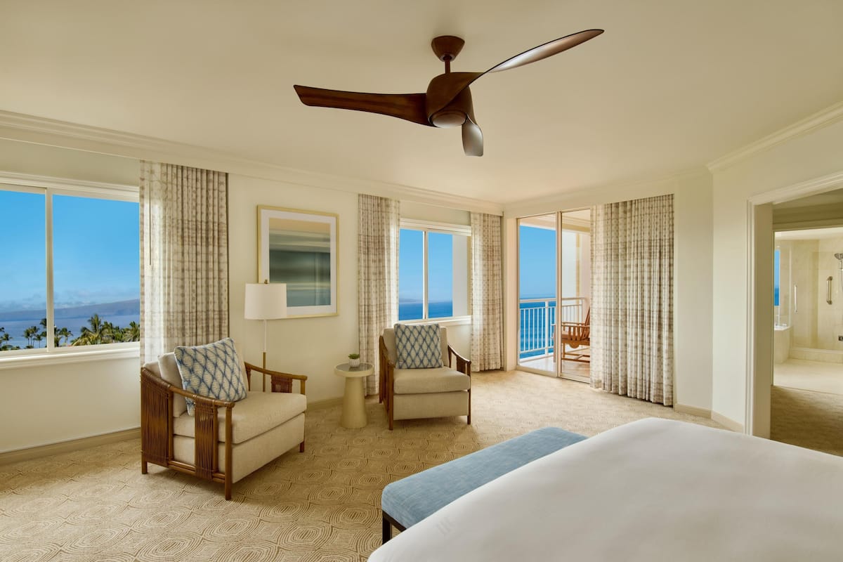 A spacious hotel room with a large bed, two chairs, a small table, and a ceiling fan.