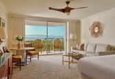 A hotel room with a balcony, a sofa, a dining table with chairs, and a ceiling fan.