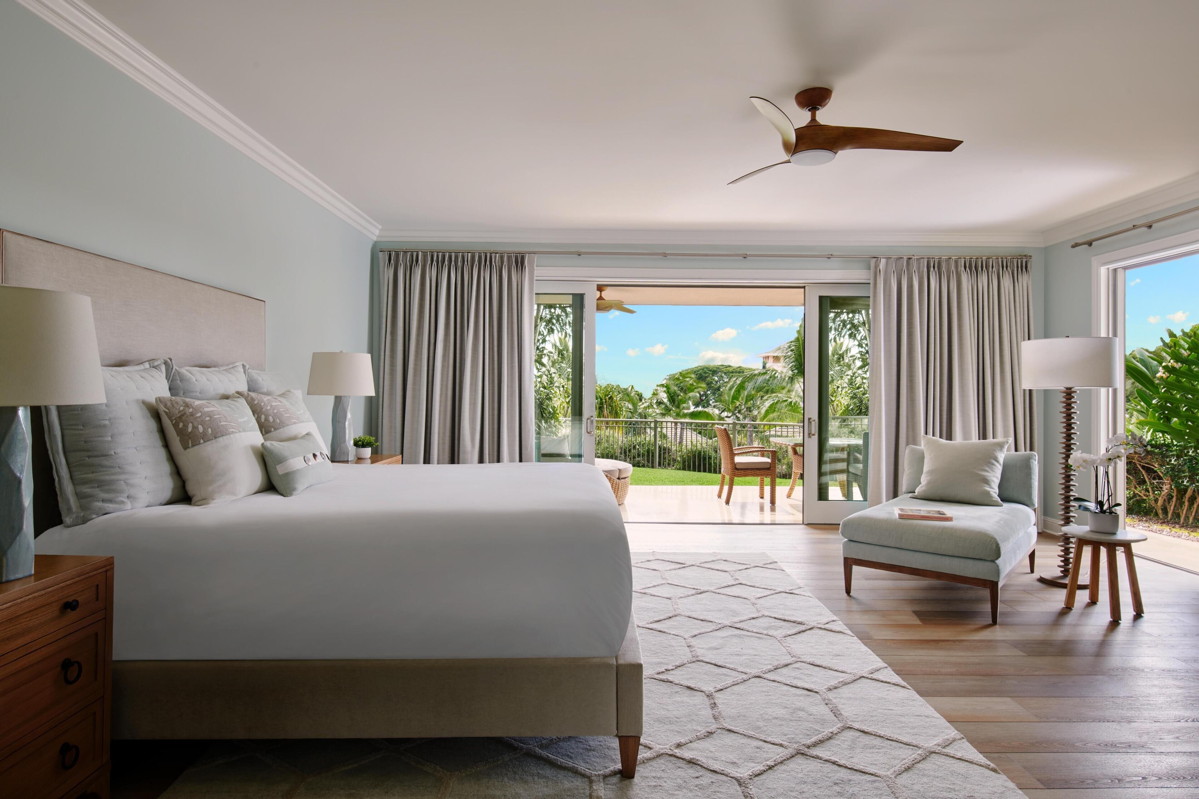 A bedroom with a large bed, a ceiling fan, a view of the outdoors, and a small seating area.
