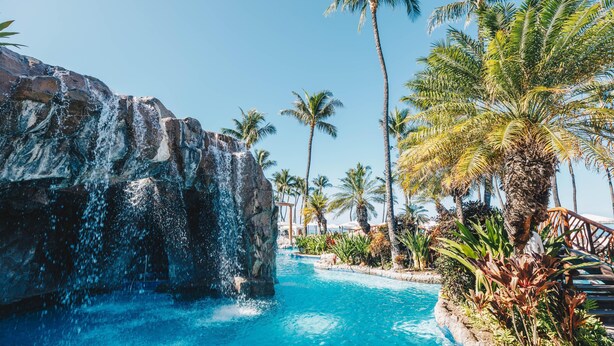 A pool with a waterfall feature, surrounded by palm trees and a stone structure. - Grand Wailea Maui, A Waldorf Astoria Resort (Kihei)