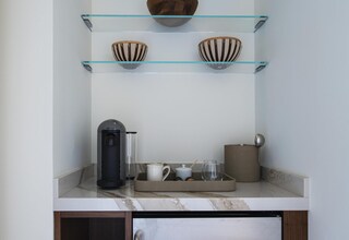 A compact kitchenette with a stainless steel refrigerator, a coffee maker, and glass shelves with decorative bowls.