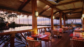 An outdoor dining area with wooden furniture and tables, overlooking a body of water and a sunset view.