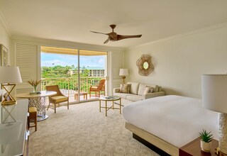 A hotel room with a bed, a desk with a chair, a small table, a sofa, a ceiling fan, and a view of the outdoors.