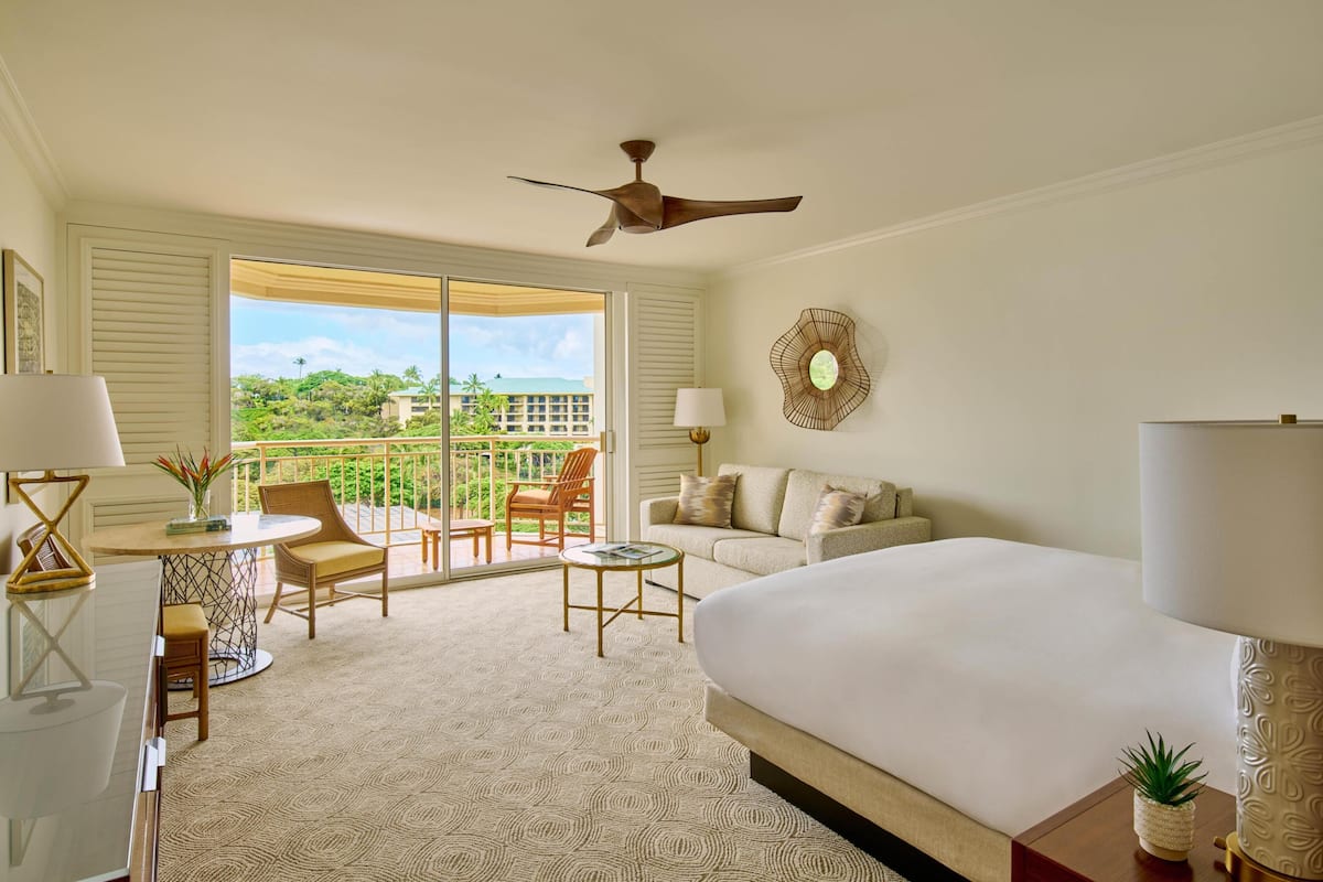 A hotel room with a bed, a desk with a chair, a small table, a sofa, a ceiling fan, and a view of the outdoors.