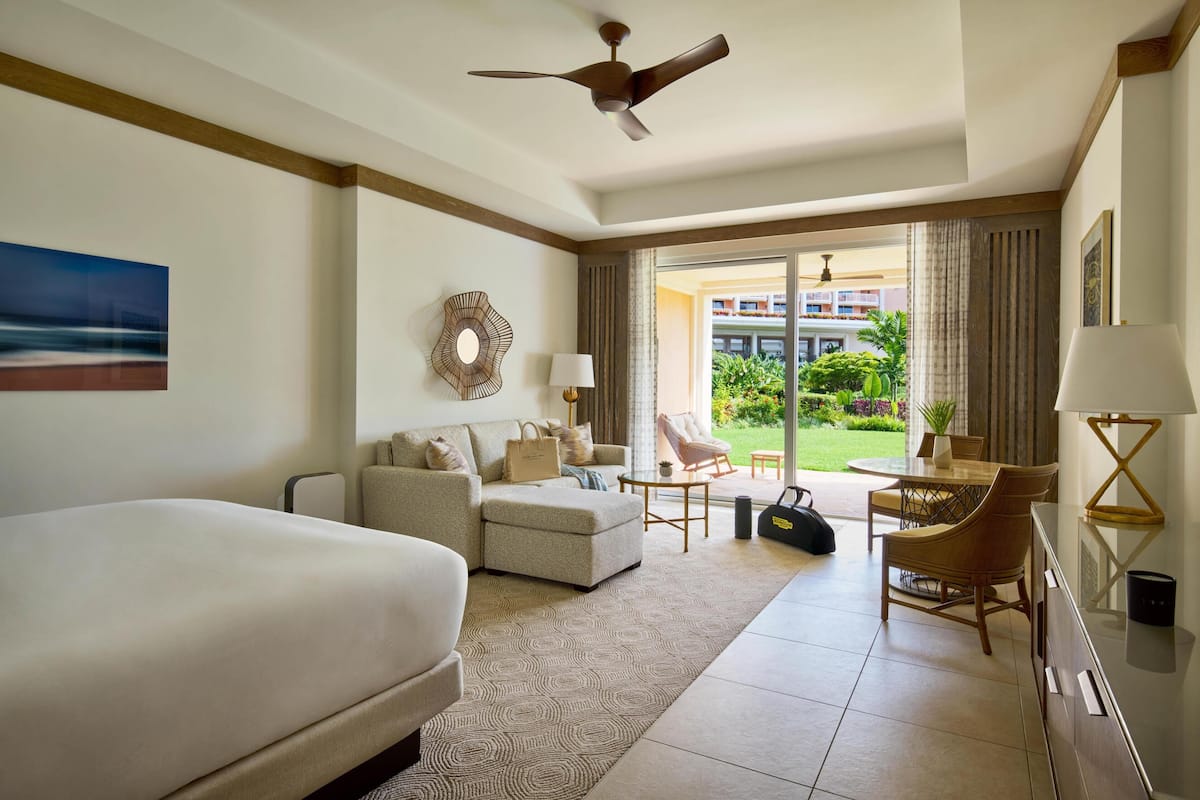A hotel room with a bed, a sofa, a dining table, a chair, a lamp, a ceiling fan, and a view of the outdoors.