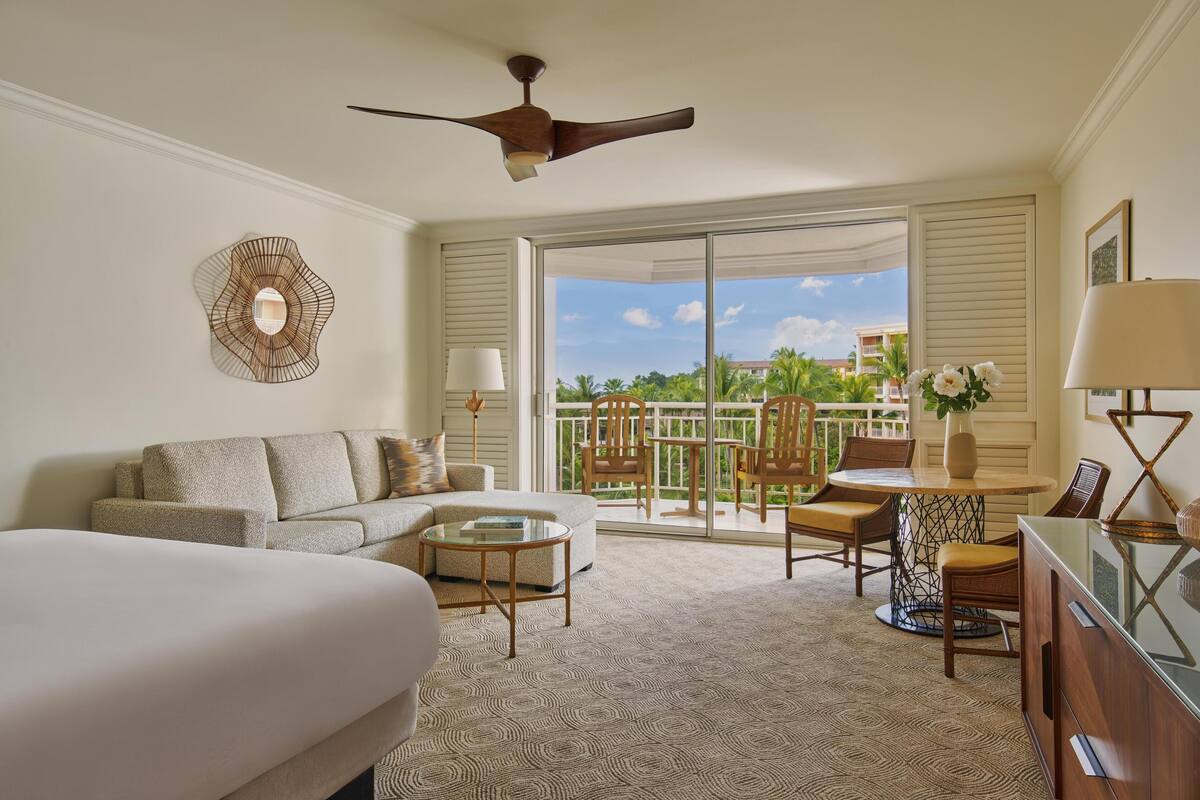 A hotel room with a bed, a sofa, a coffee table, a dining table with chairs, a ceiling fan, and a balcony with a view of palm trees and buildings.