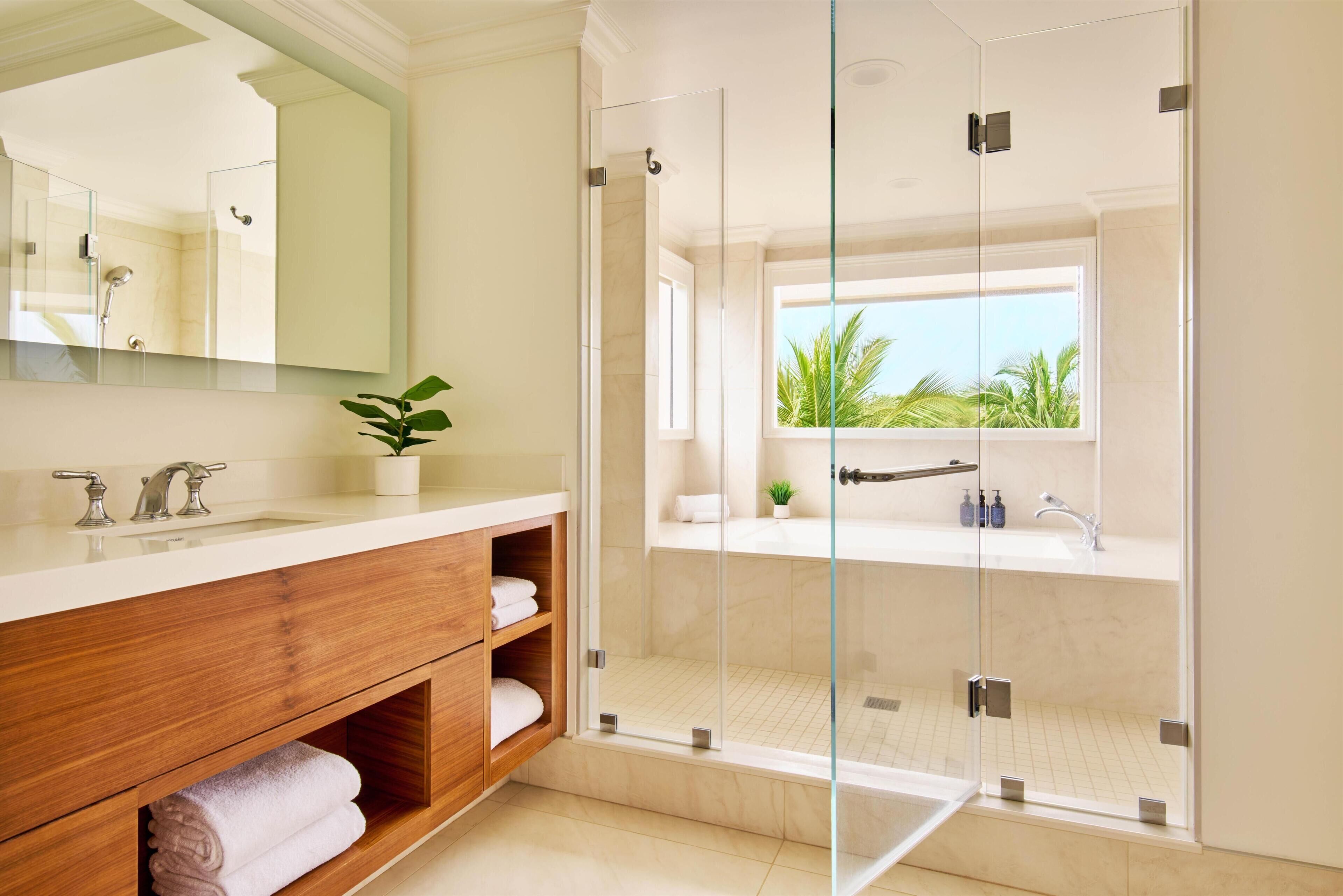 Un baño moderno con ducha de vidrio, tocador de madera, un espejo grande y vistas a palmeras.