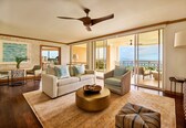 A spacious living room with a sofa, armchair, and round coffee table, a ceiling fan, and a balcony with a view.