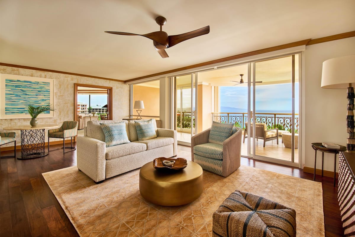 A spacious living room with a sofa, armchair, and round coffee table, a ceiling fan, and a balcony with a view.