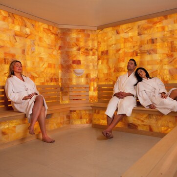 Three people in white robes sitting on wooden benches in a room with mosaic walls.