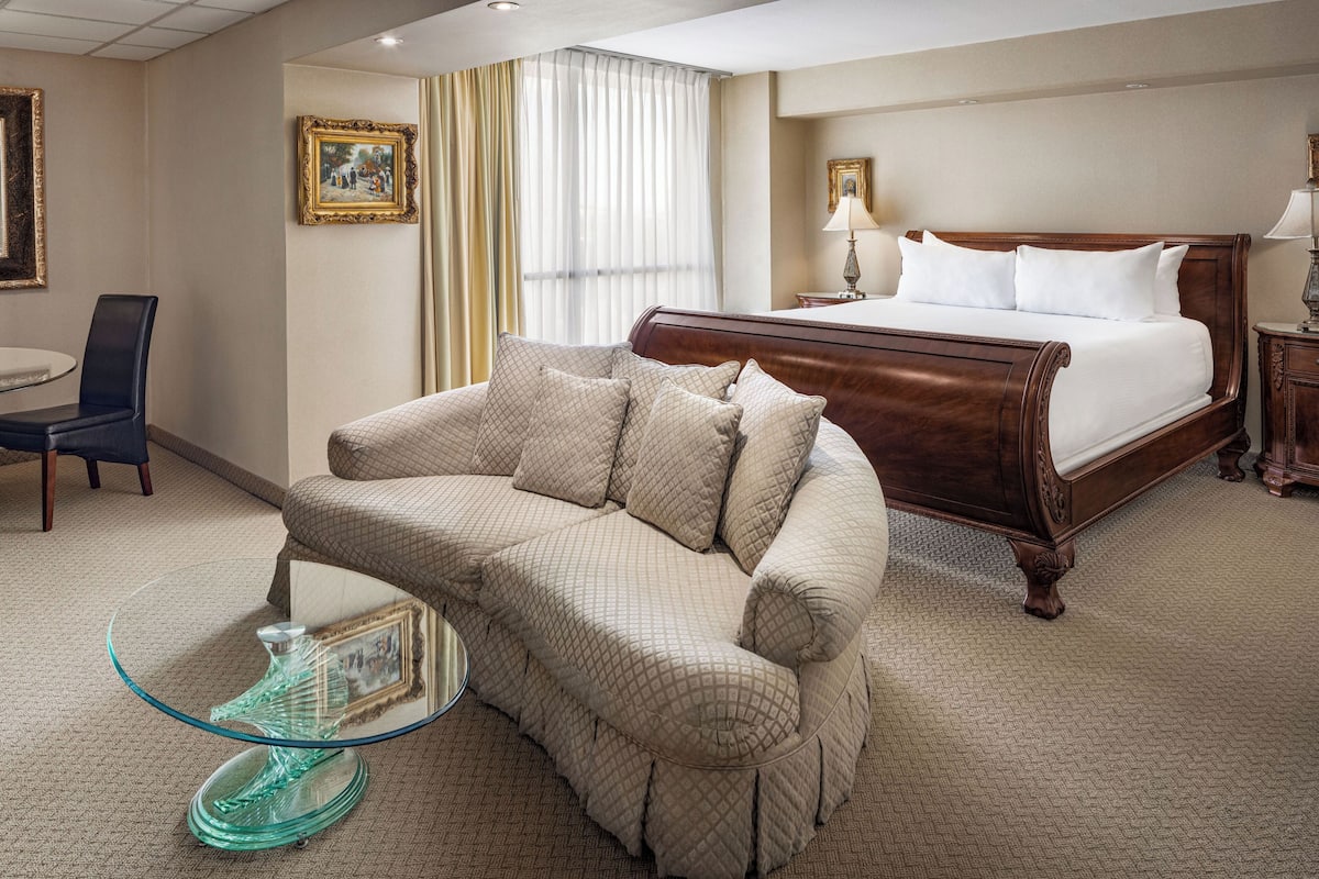 A hotel room with a large bed, a sofa, a glass coffee table, and a dining area with a chair and table.