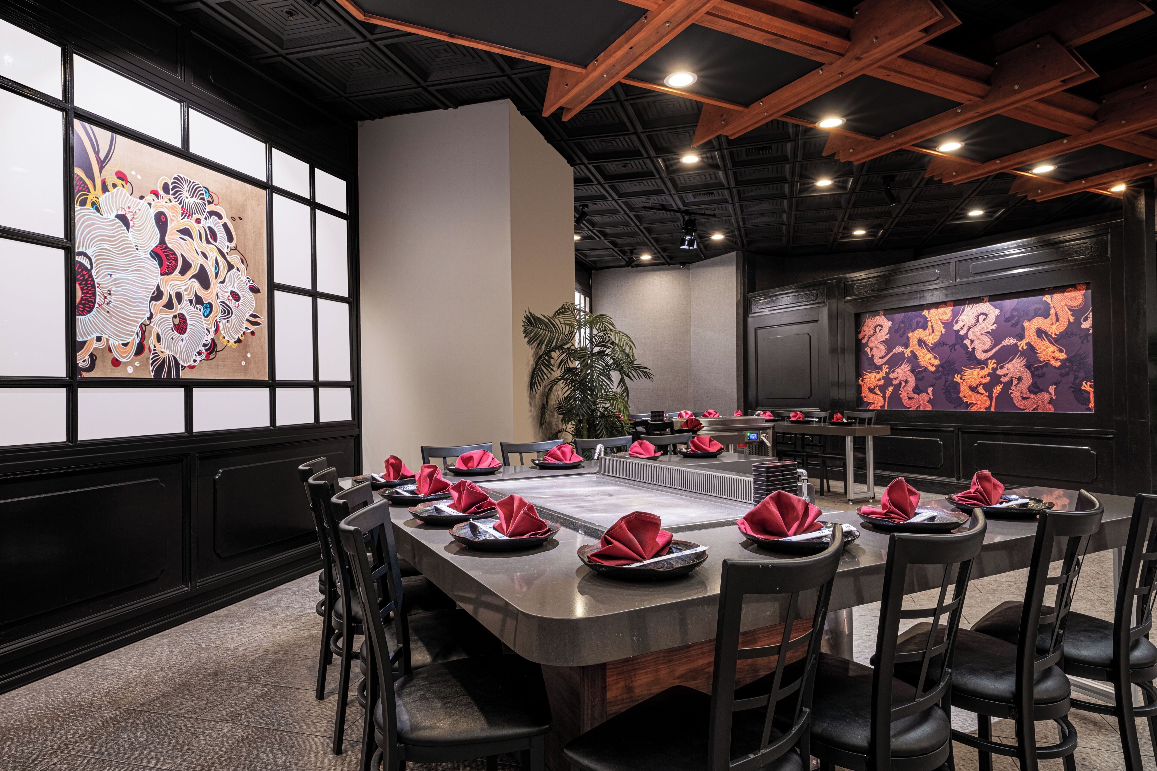 A restaurant with a long communal table, black chairs, and red napkins.