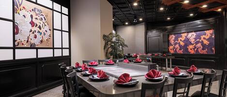 A restaurant with a long communal table, black chairs, and red napkins.