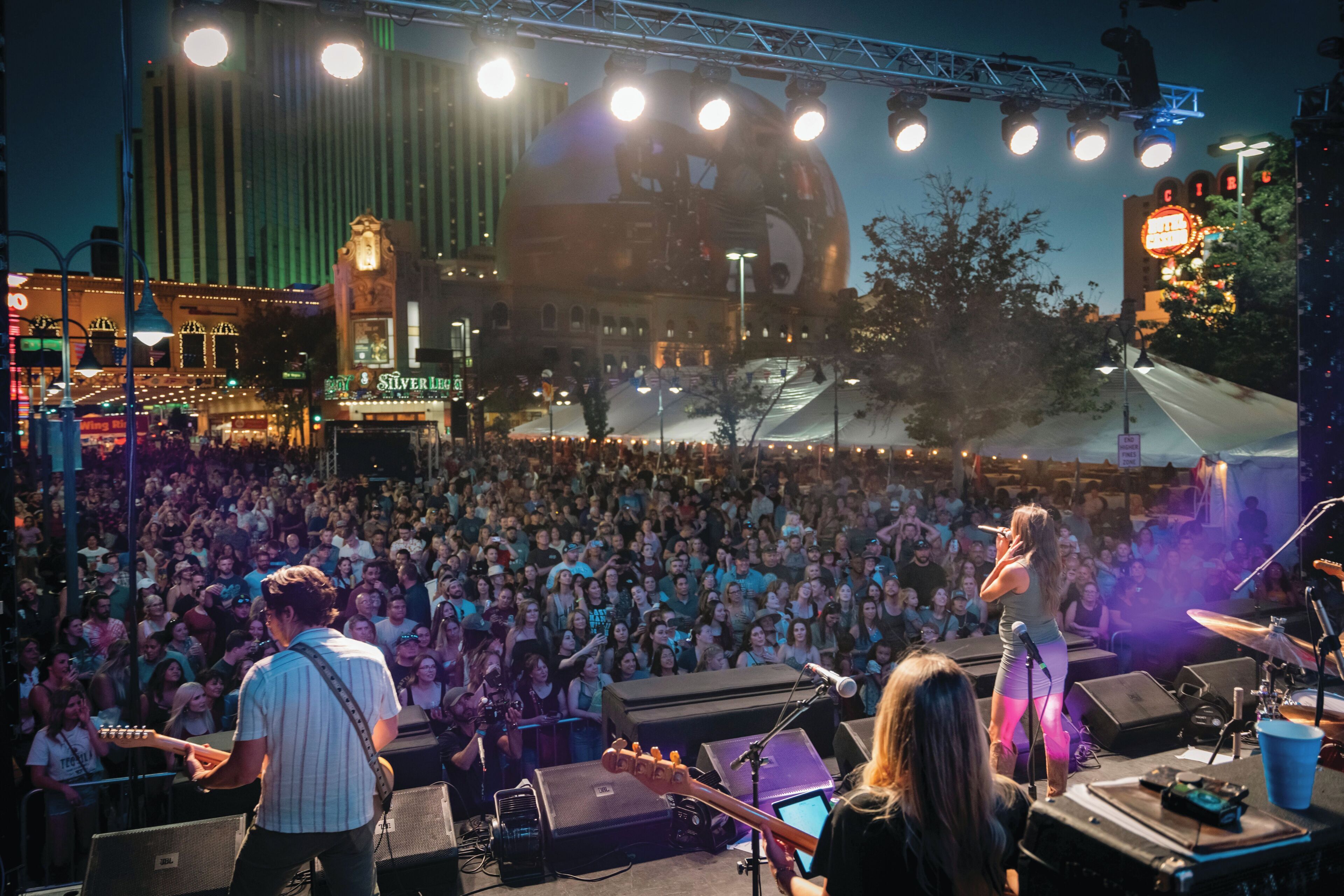A concert stage with musicians performing, a large audience, and a city skyline in the background.