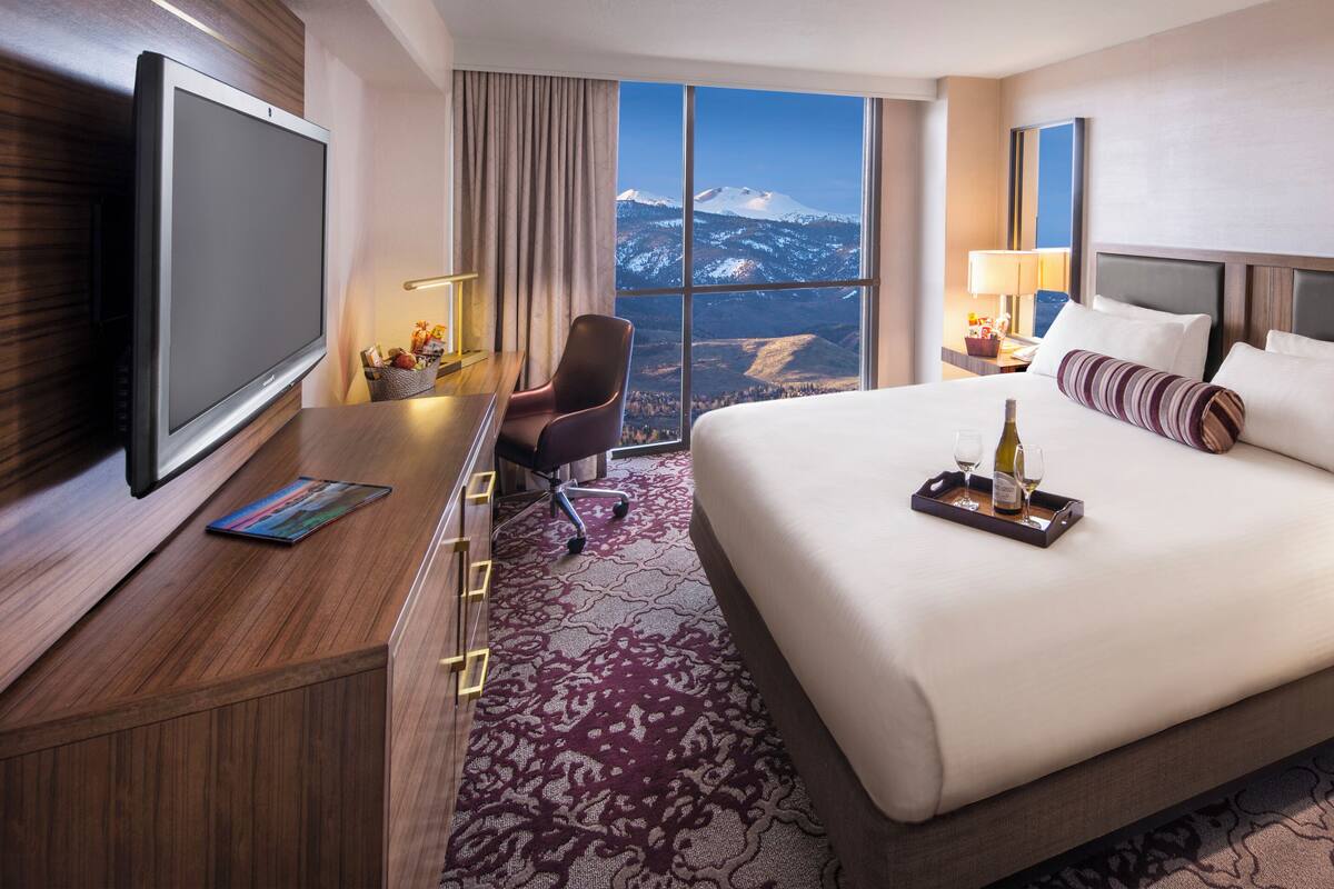 A hotel room with a large bed, a desk with a flat-screen TV, a chair, a nightstand with a lamp, and a view of snow-capped mountains.