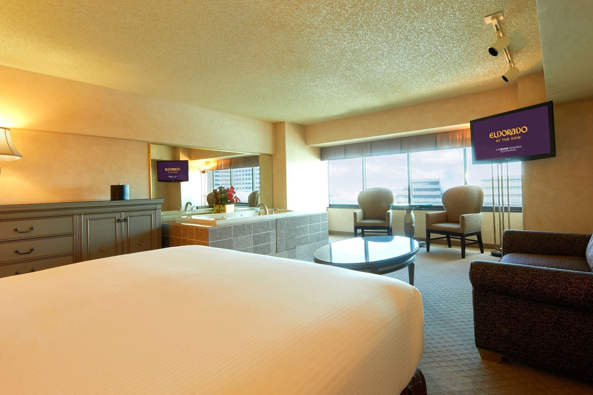 A hotel room with a large bed, a desk with a lamp, a TV, a sofa, and a view of the city.