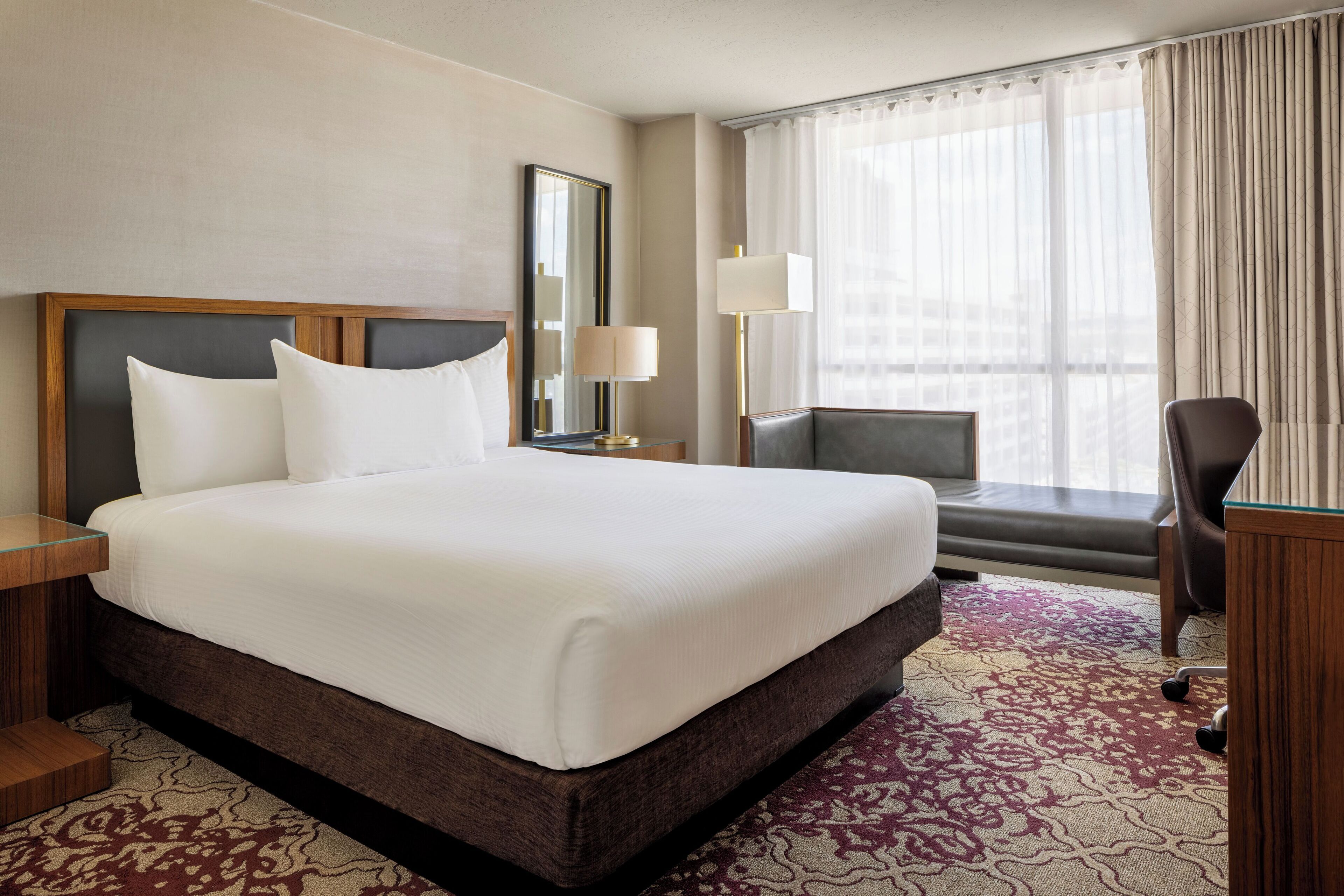 A hotel room with a large bed, a desk, a chair, a window with curtains, and a nightstand with a lamp.