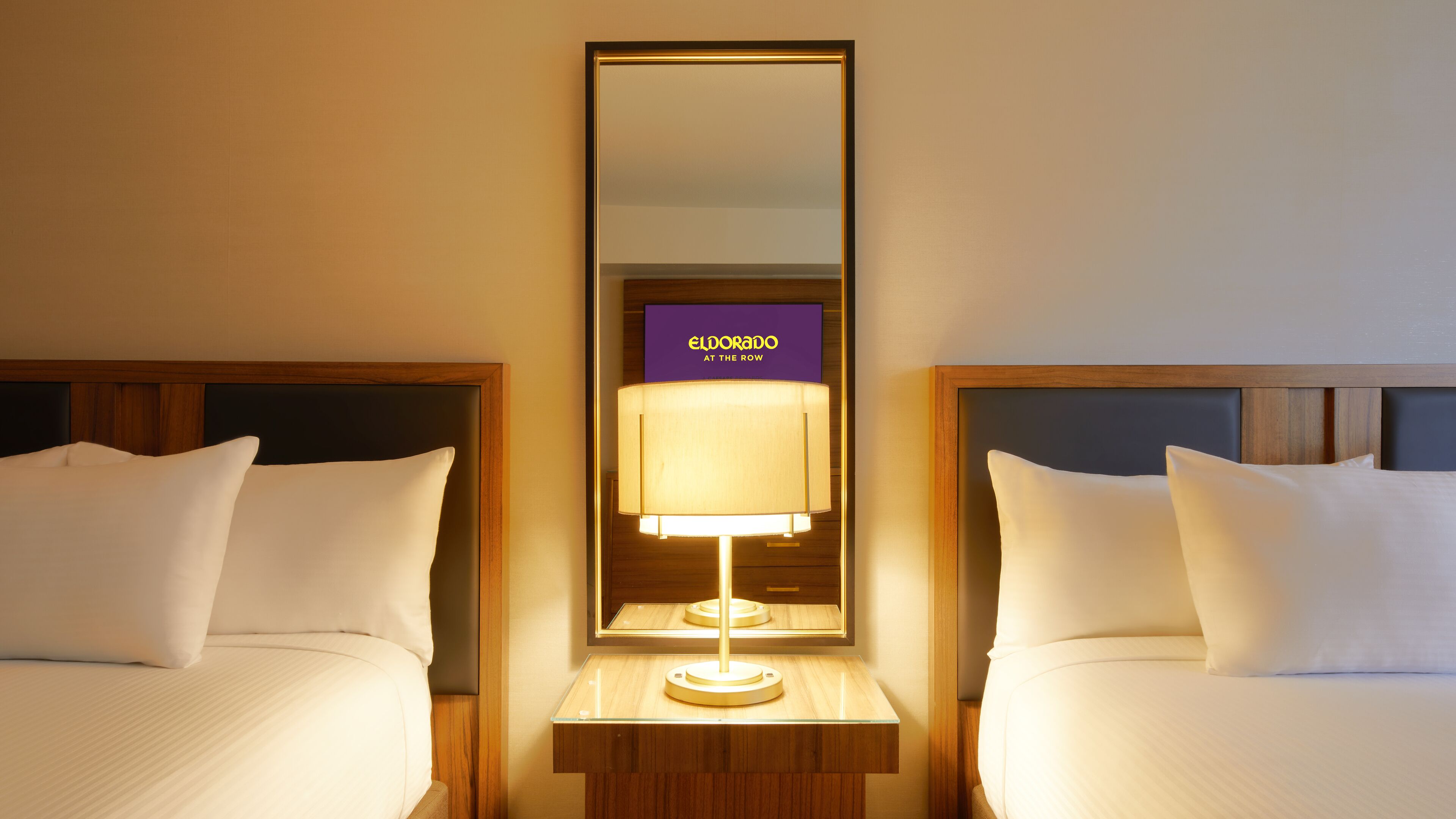 A hotel room with two beds, a nightstand with a lamp, and a mirror reflecting the room.