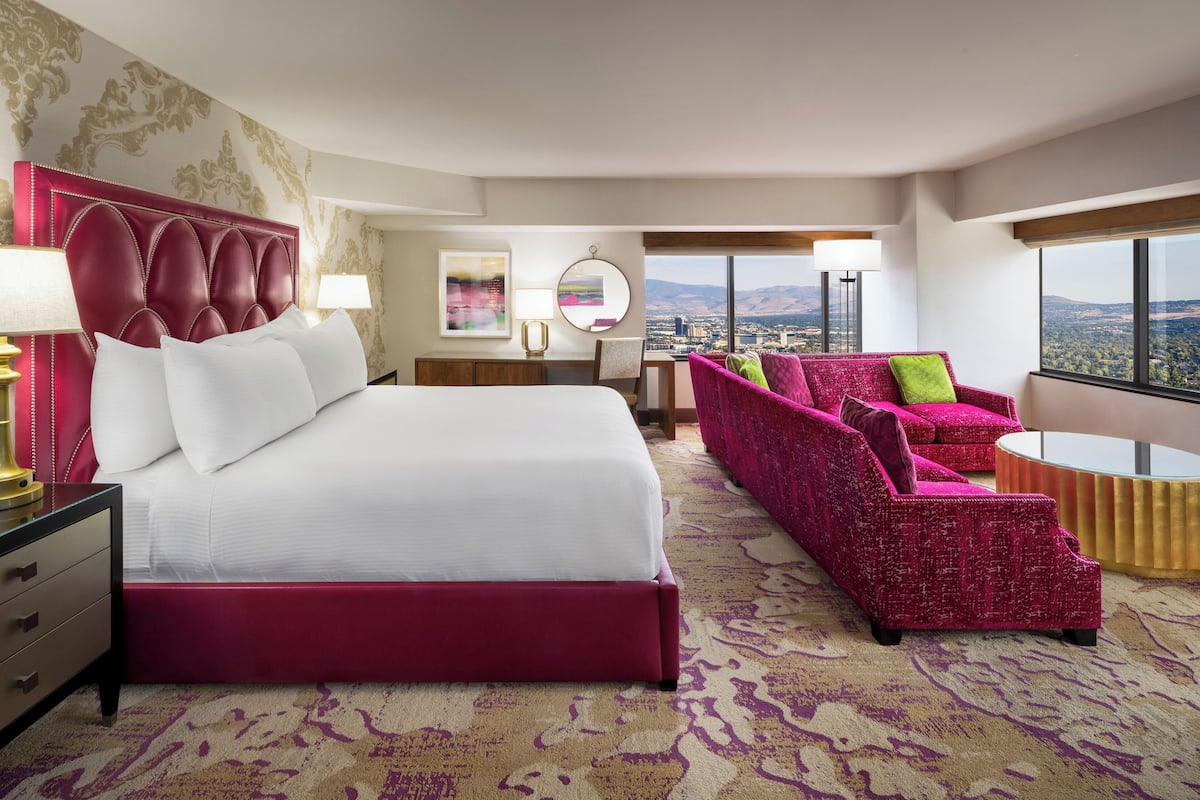 A hotel room with a large bed, a pink sofa, a desk, and a view of the city.