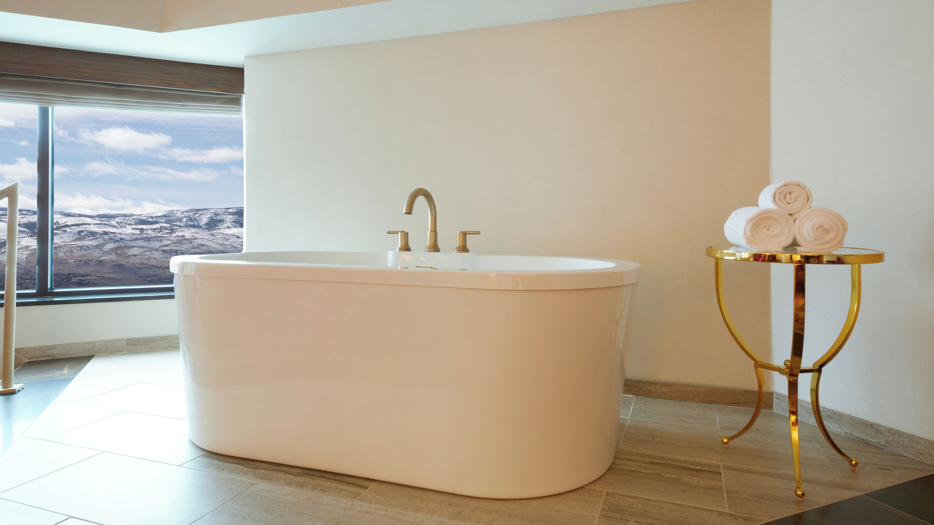 Une salle de bain moderne avec une baignoire sur pieds, une coiffeuse aux montures dorées agrémentée de serviettes et une grande fenêtre offrant une vue panoramique.