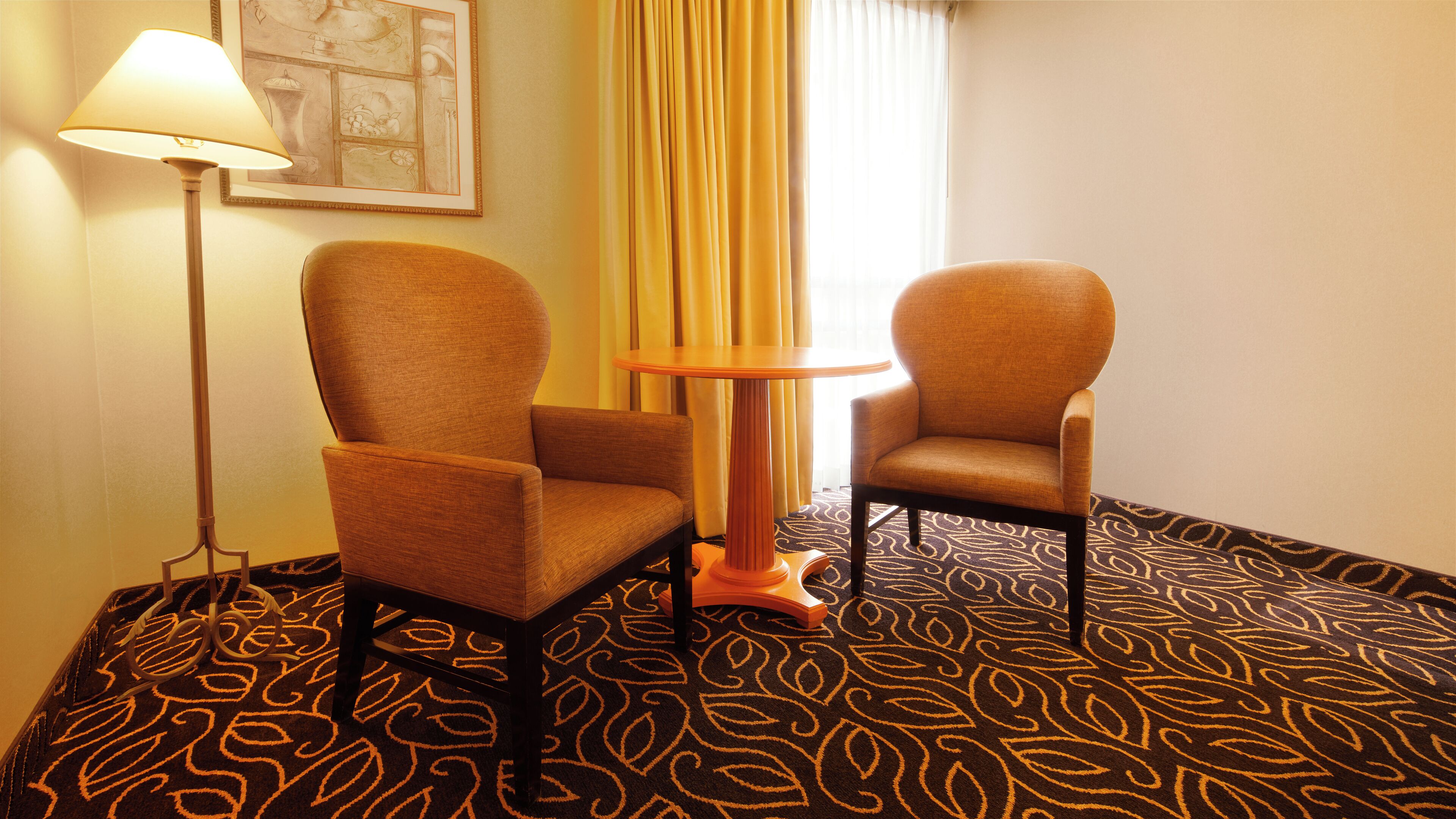 A hotel room with two armchairs, a small round table, a floor lamp, and a patterned carpet.
