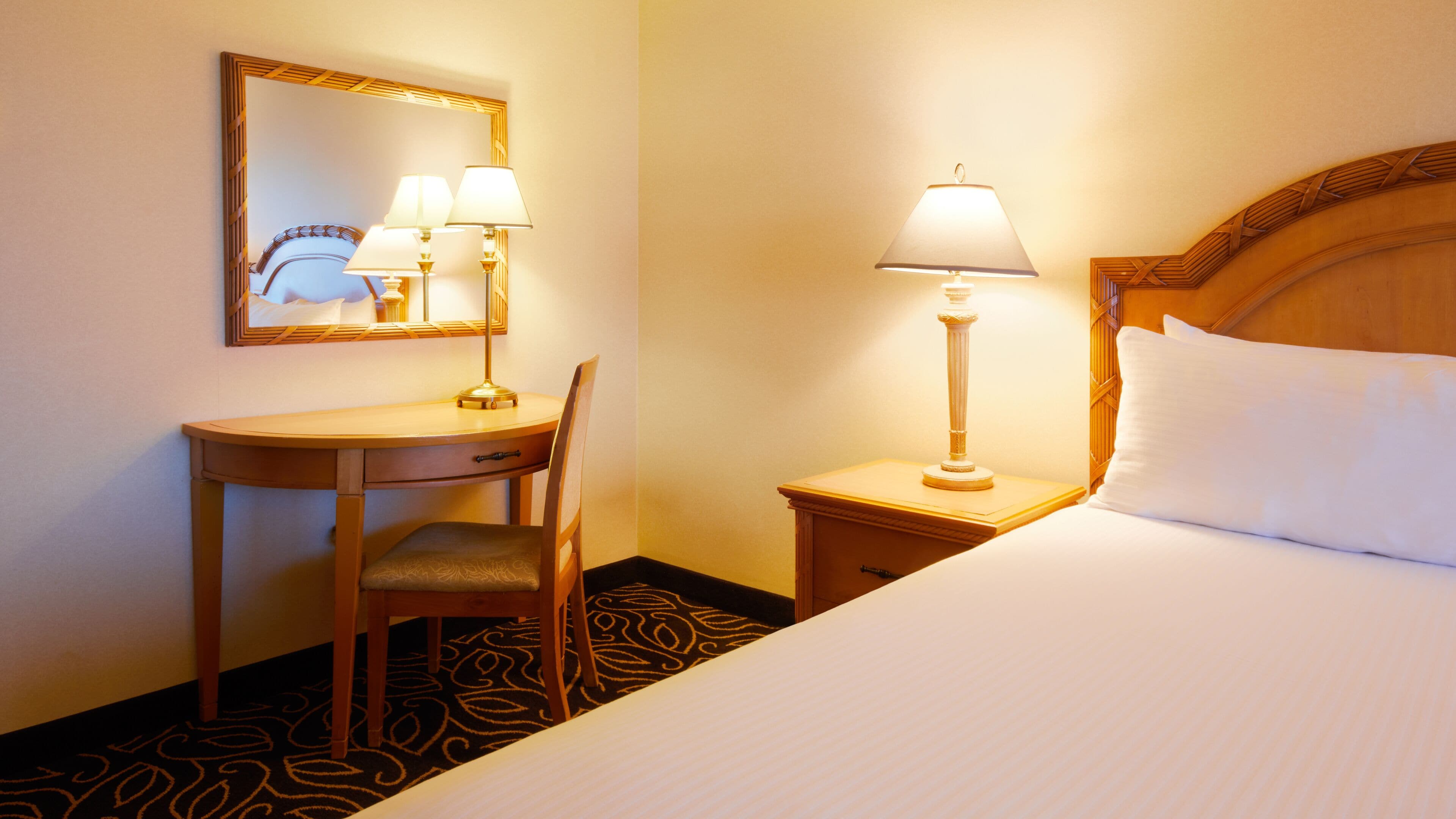 Habitación de hotel con cama, escritorio con silla, mesita de noche con lámpara y espejo en la pared.