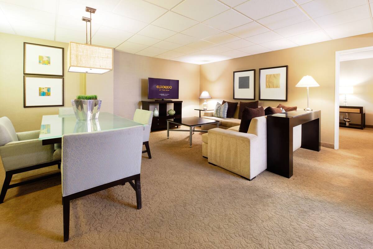 A modern hotel room with a glass dining table, comfortable seating, a flat-screen TV, and a cozy seating area with a sofa and armchairs.