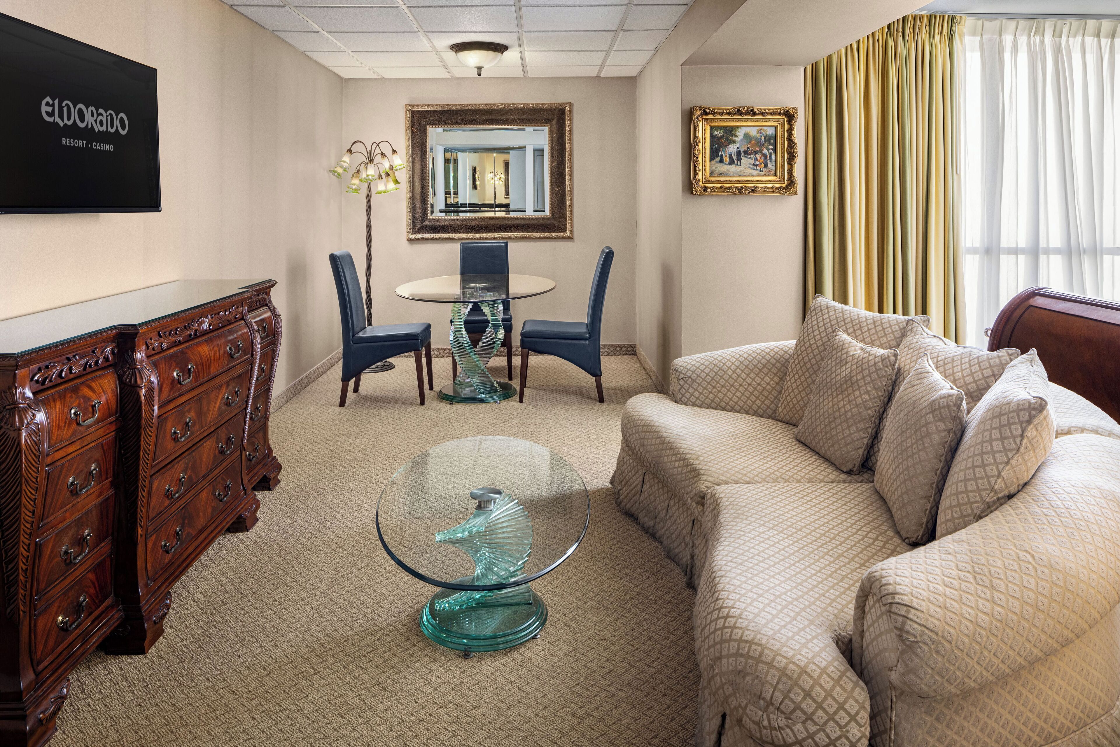 A hotel room with a wooden dresser, a glass coffee table, a beige sofa, a dining table with chairs, a mirror, and a framed picture on the wall.